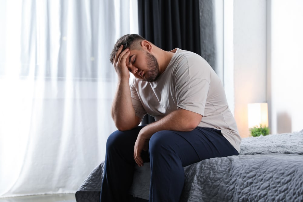 A bearded man struggling with his weight and sleep apnea is sitting on a gray bed and rubbing his head while is eyes are closed.