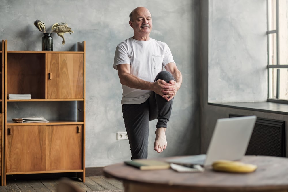 An older man suffering from sleep apnea is trying to lose weight by working out and eating better. He's standing in front of a laptop and is pulling his left knee up toward his chest. There's a banana behind his laptop.