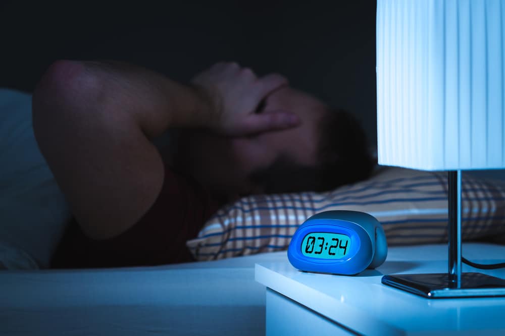 A man suffering from sleep apnea is awake late at night. His alarm clock reads 3:24 am and his hand is over his face.