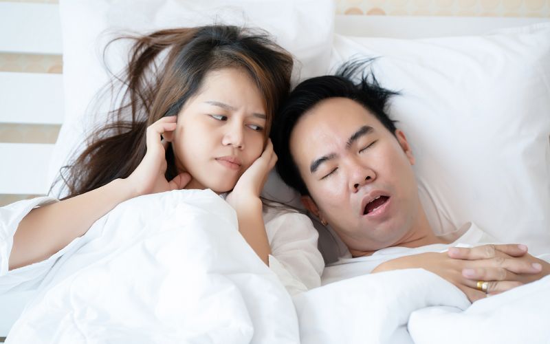 a couple in bed together with the man snoring and the woman covering her hears while glaring